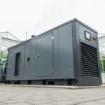 Large industrial diesel generator installed at a cannabis production facility with greenhouse buildings blurred in the background, conveying reliable 24/7 power readiness.